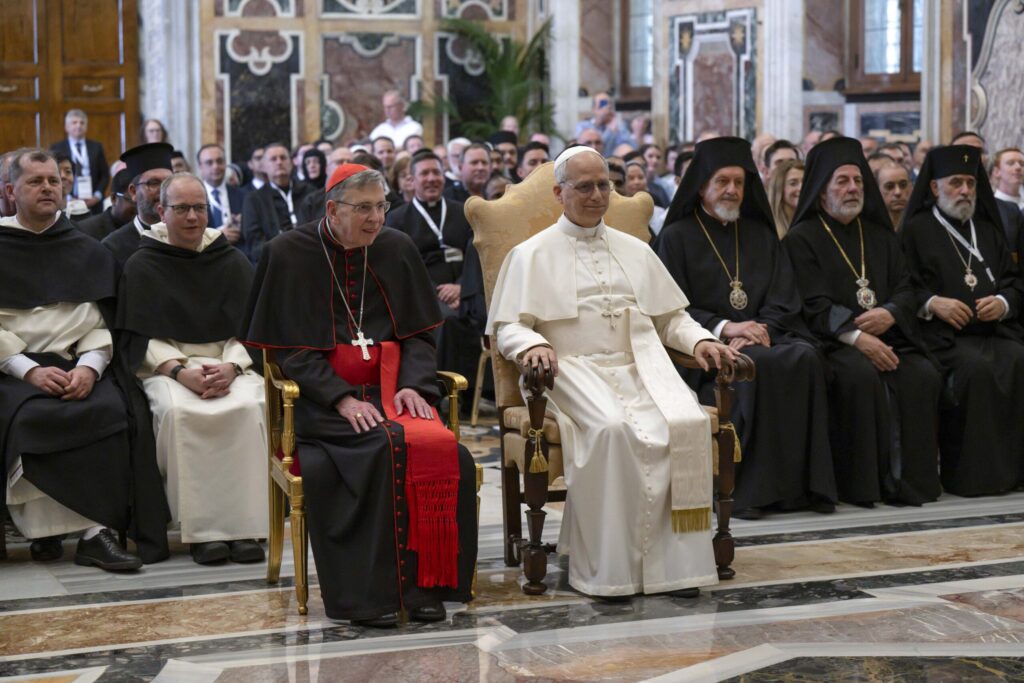 Pope Leo XIV sits in a Vatican conference hall next to Cardinal Kurt Koch, surrounded by clergy from the Roman Catholic and Orthodox Churches.