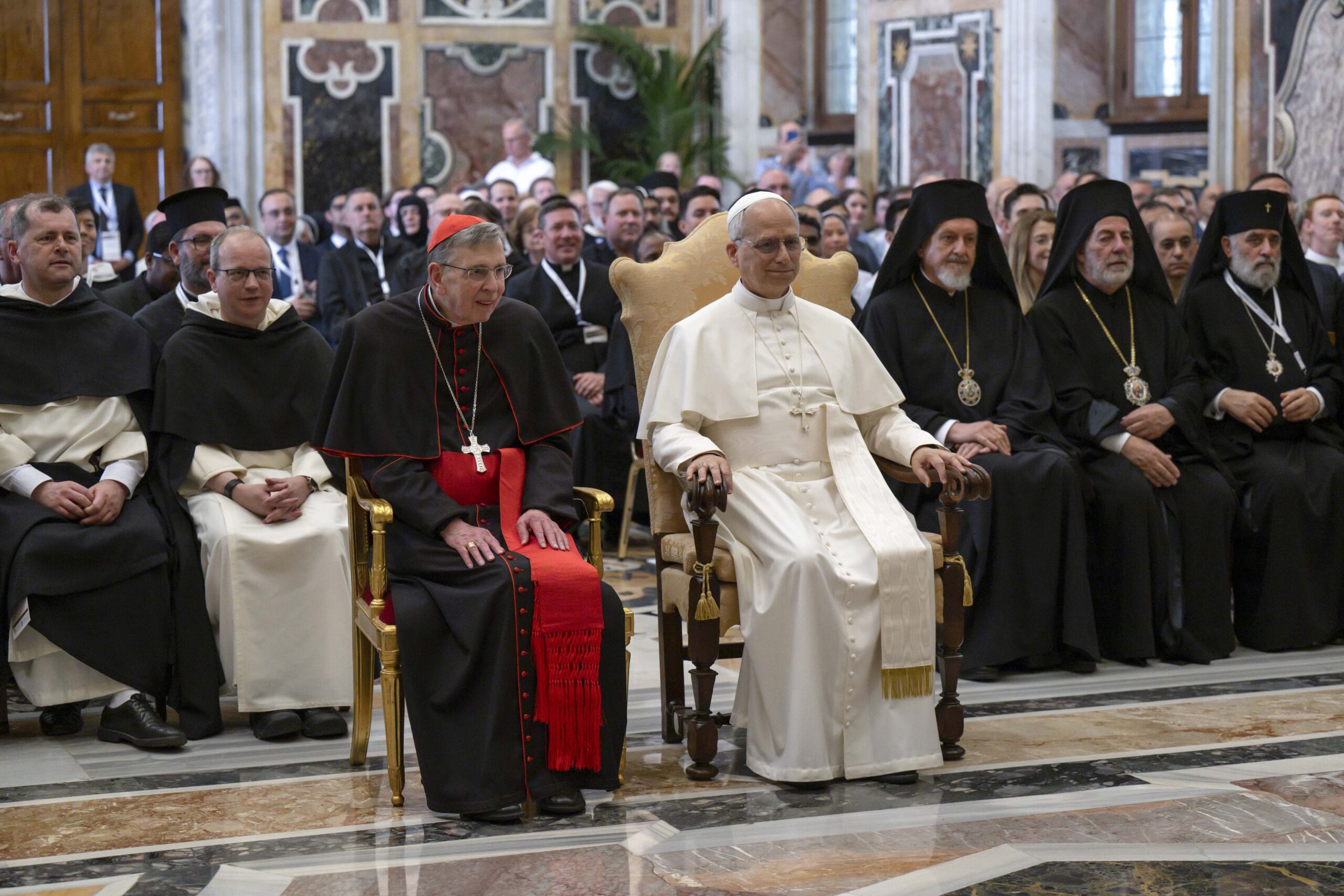Pope Leo XIV sits in a Vatican conference hall next to Cardinal Kurt Koch, surrounded by clergy from the Roman Catholic and Orthodox Churches.